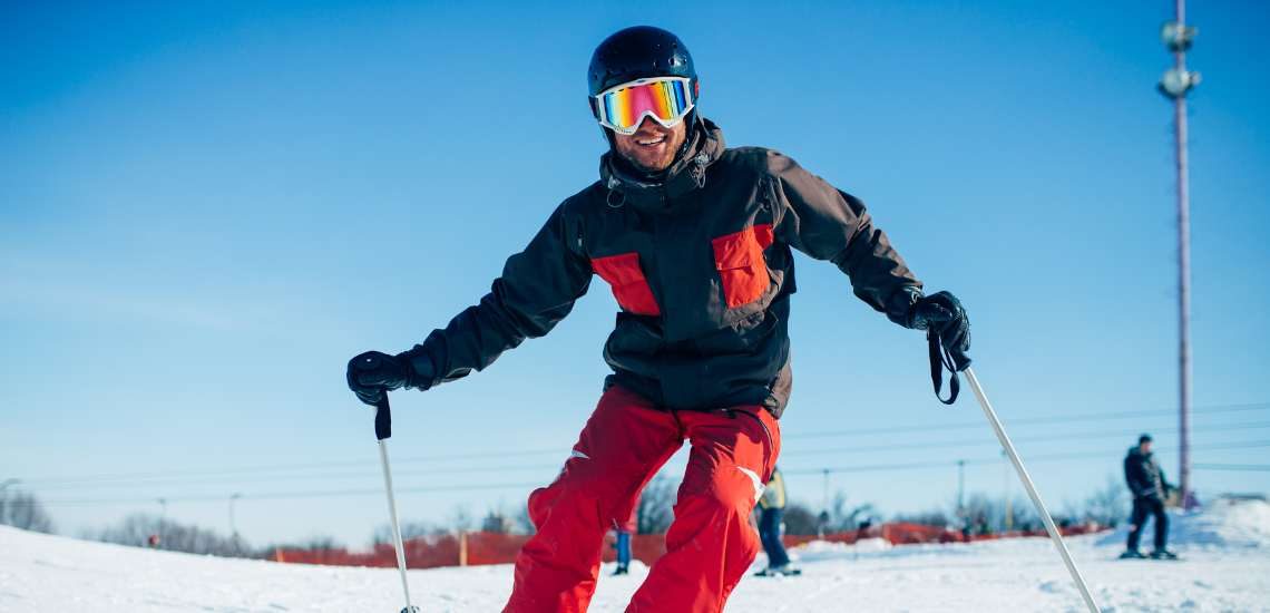 Ein grinsender Mann mit einem Ski-Anzug und Ski-Helm fährt einen Hang hinunter.